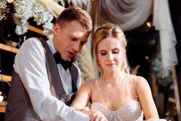 Naklejka premium A groom in a tuxedo and a bride in a dress cut the wedding cake at the end of the evening. Wedding traditions of the couple. Emotions and joy from starting a family.