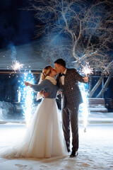 A groom in a jacket and a bride in a jacket are holding sparklers and fireworks in their hands. Kisses against the background of cold sparkling fountains outside in winter. 