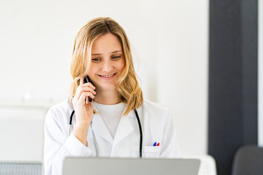 Smiling blond female doctor talking on smart phone in medical clinic