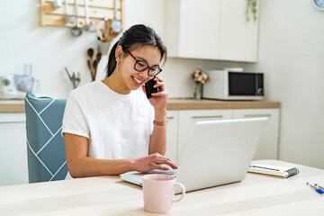 Young woman talking on mobile phone while using laptop in kitchen