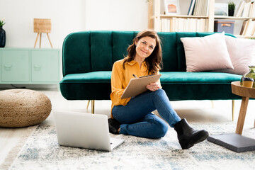 Smiling mature woman writing on clipboard while sitting at sofa in living room
