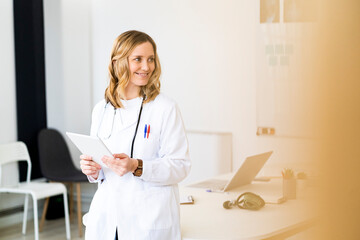 Smiling female healthcare worker with tablet leaning on desk while standing in medical clinic