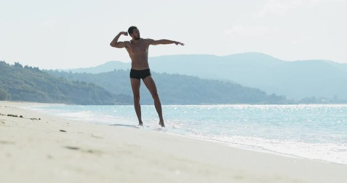 The handsome man with a perfect athletic body in swimming trunks having fun on a deserted beach in the morning, He brags of the body as the bodybuilder, showing off his body and posing