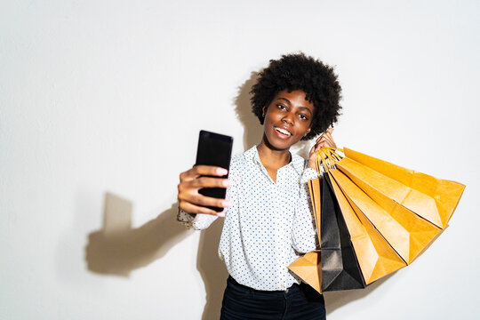 Young smiling woman with shopping bags taking selfie through smart phone while standing against white background