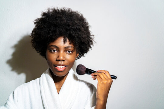 Curly woman using blushbrush while standing against white background