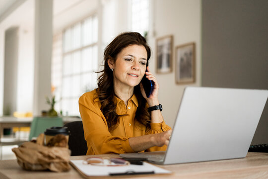 Smiling businesswoman using laptop while talking on mobile phone at desk in home office