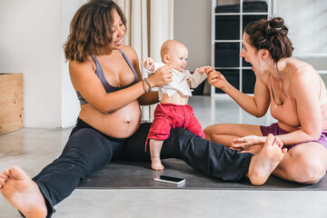 Two young women in the gym after pilates workout have fun and talk - Mother with baby and pregnant person sitting on mat relax after training - Concept of motherhood and family