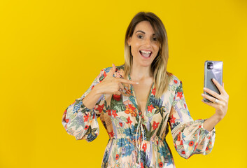 Smiling woman in summer dress using mobile phone with surprised face isolated over yellow background.