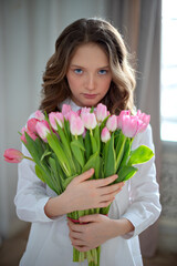 A beautiful little girl in a white shirt with long curly brown hair and blue eyes holds a huge bouquet of Tulips in her hands