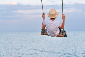 Boy rides on a swing over the water back view. Vertical frame.