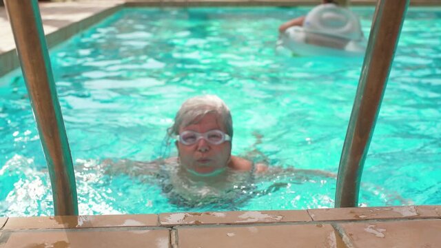 Elderly senior woman with gray hair swimming in hotel pool, googles on eyes, smiling satisfied as she gets near