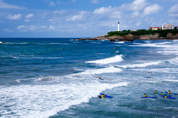Surf school students in the middle of blue ocean waves, aerial panorama view, white lighthouse tower on the rocky hill behind 