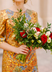 A bride in an unusual golden dress holds a bridal bouquet of white and red roses in her hands, close-up 