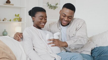 African American couple family future expecting parents sitting on couch afro black husband man stroking wife pregnant belly with cute small childish child white shoes hugging embracing beloved woman