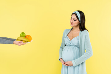 smiling pregnant woman touching belly while looking at fresh fruits isolated on yellow.