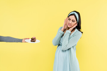 pregnant woman feeling disgust near piece of delicious cake isolated on yellow.