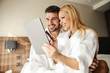 Man and a woman with a tablet and a credit card. They make online reservations in the hotel room via the wireless internet in the comfort of a bathrobe. Cuddling in bed, sharing loving memories