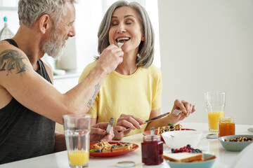 Older husband feeding mature wife tasting waffles having breakfast at home. Happy healthy...