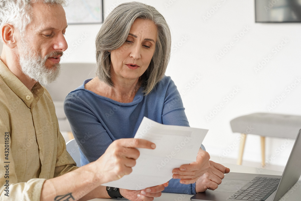 Wall mural Older mature couple checking bank documents using laptop at home. Senior mid age retired man and woman reading paper bills, calculating pension or taxes, planning retirement finances, doing paperwork.