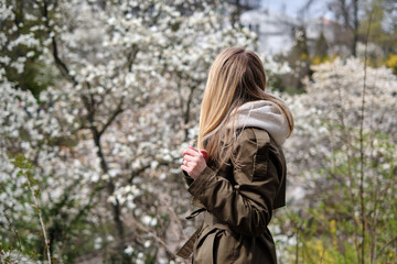 Fototapeta na wymiar Beautiful young girl walks among the flowering trees of magnolia in the park. Happy woman enjoys flowers flavors and spring mood. White blooming trees. Spring season concept. Walk outdoors