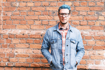 Stylish and fashionable handsome young man with blue hair wearing in jeans jacket standing on background of brick wall. Model looks directly into the camera and smiles slightly.
