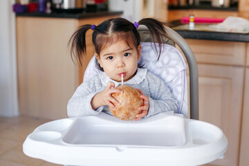  Cute adorable Asian Chinese kid girl sitting in high chair eating soup with spoon. Healthy eating for kids children. Toddler eating independently in kitchen at home. Candid real authentic moment.