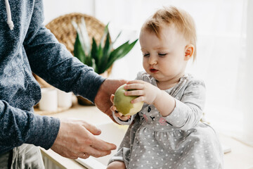little red haired girl is sitting in the kitchen of a country house eating an apple from father's hands