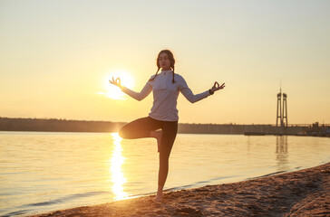 Fototapeta premium Young woman meditating doing yoga on sandy beach by the river on background of setting sun