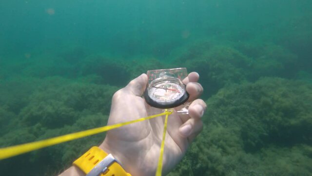 First-person view of a male hand holding a plastic magnetic compass rotates from the side and seeks the correct direction underwater.