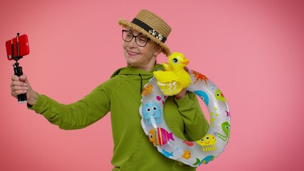 Mature grandmother traveler blogger with swimming ring and inflatable duck toy taking selfie on mobile phone make video call online. Studio shot. Senior woman on summer holiday vacation, trip. Tourism