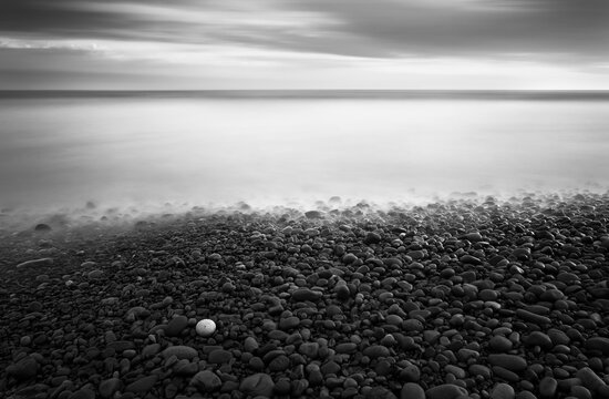 A black and white image of one yellow pebble that stands out against all other black and grey pebbles in a fine art long exposure of the ocean and clouds.
