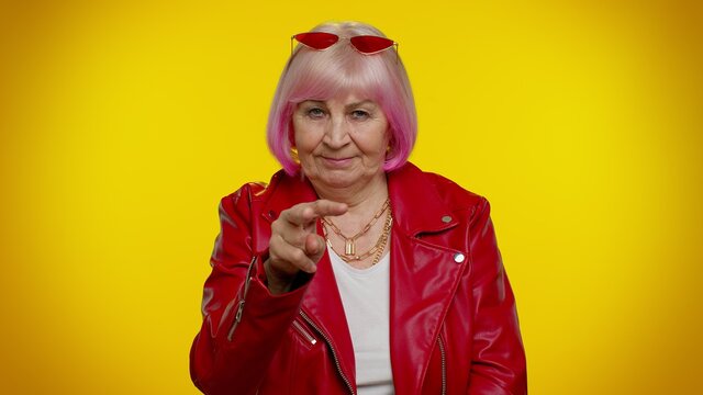 Elderly Rocker Granny Woman Pointing At Her Eyes And Camera, Show I Am Watching You Gesture, Spying On Someone. Senior Old Grandmother Isolated On Yellow Background. People Lifestyle Emotions