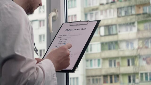 Young doctor fills in patient information, near the window, against the background of the building, therapy and doctor concept