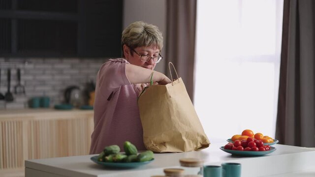 grandma is unpacking food after shopping in market at weekend, taking fresh greenery and bread, medium portrait of single elderly lady at home