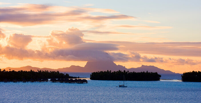 French Polynesia: Three small islets with main island in background at sunset with orange and purple clouds