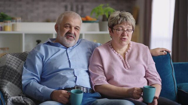 grey haired man is embracing his aged wife in living room, spouses are sitting together in living room and spending time at weekend