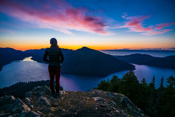 Adventurous athletic woman hiker standing on a mountain top watching the sunset over mountains and a beautiful lake.