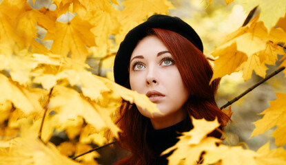 Naklejka na ściany i meble Cute girl in yellow foliage looks up. The fall period trees. Autumn romantic portrait of a red-haired woman. Walk in the Park.