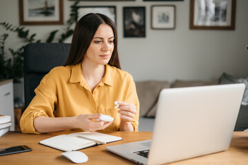 Young pretty woman talking on video call and waving hand while sitting at table in the homeoffice room