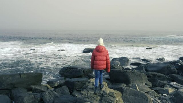Back view. A girl in a red jacket slowly walks over the stones to the seashore. The child jumps from stone to stone, approaching the ocean.