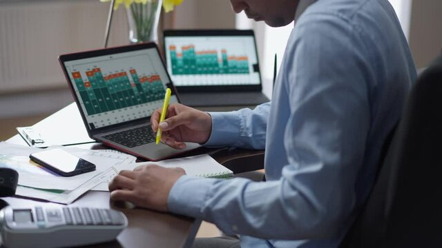 Unrecognizable African American man analyzing graphs on laptops screens and writing down notes. Serious confident analyst working remotely in home office indoors. Business efficiency concept