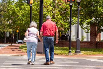 Senior caucasian couple in love holding hands as they cross the street on pedestrian crossing on a lovely spring day. Activity, togetherness at elderly community concept. Woman carries a sidebag.