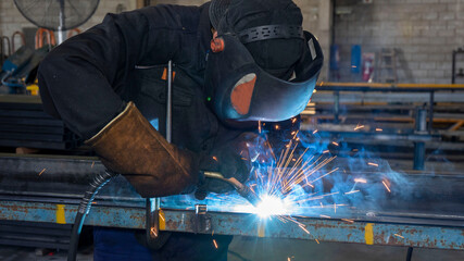 Worker welding in steel workshop with bright light and sparks.