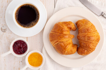 Flat lay plain soft croissant bread on a white plate served with a cup of coffee, orange spread and mixed berry spread for breakfast or coffee break time.