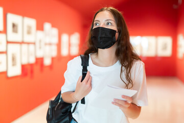 Focused girl visitor in a protective mask admires the exhibition of paintings, holding a paper guide to the museum