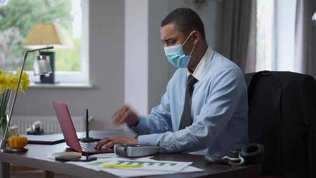 Young focused African American man in coronavirus face mask coughing messaging online and using label maker. Side view of concentrated professional manager with symptoms of Covid-19 in home office