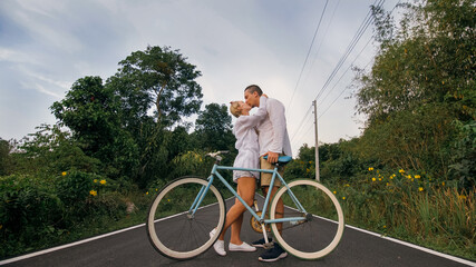 Biking road trip. Love couple on blue bike in white clothes on forest road. Just married woman and man kiss, hugs, stand on bicycle. Cycle Fix.