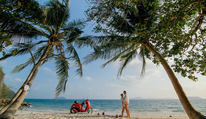 Scooter road trip. Lovely couple on red motorbike in white clothes on sand beach. People walking near the tropical palm trees, sea. Motorcycle rent.