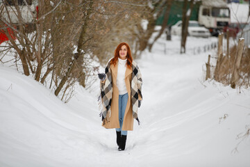 Happy woman wearing beige coat and large plaid scarf. Romantic walk in winter snow park in rustic style. Female in casual classic clothes with red hairs. Redhead smiling young girl with dried flowers