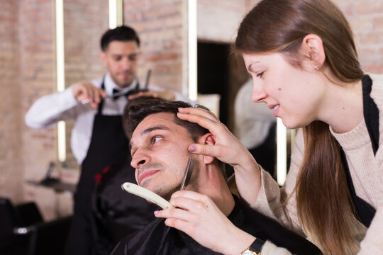 Male client getting old-fashioned shave with straight razor from female barber in modern salon.